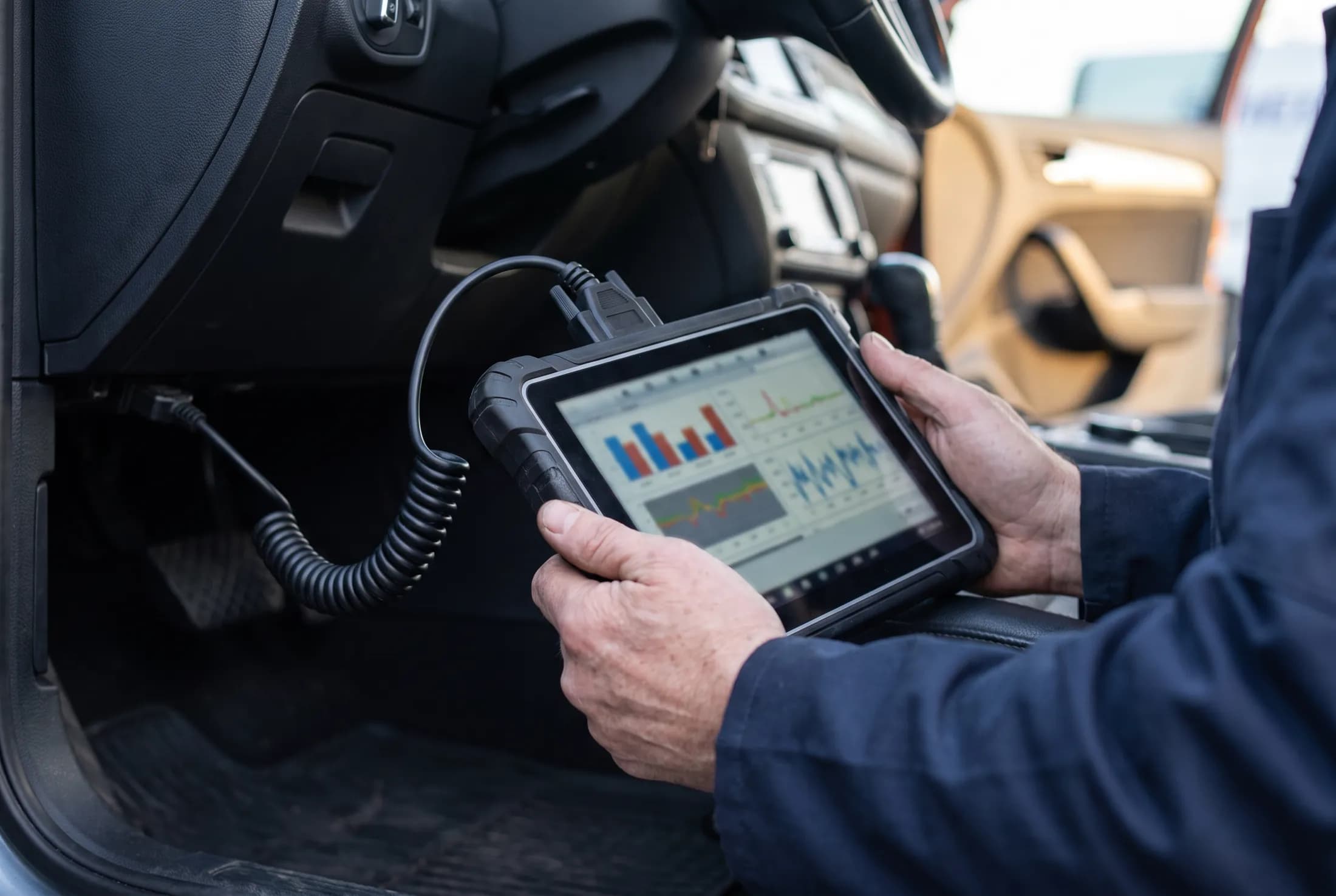 Hände eines Mechanikers mit einem Diagnosegerät, das über ein Kabel mit der OBD-Schnittstelle eines Fahrzeugs verbunden ist